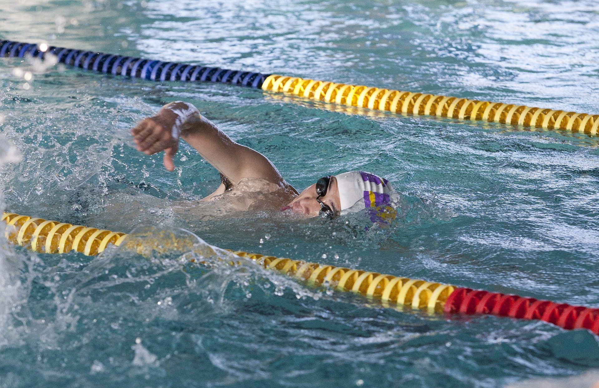 Hombre nadando en piscina olímpica. La persona tiene lentes para nadar negros y un gorro para piscina blanco con líneas moradas y amarillas a los lados. 