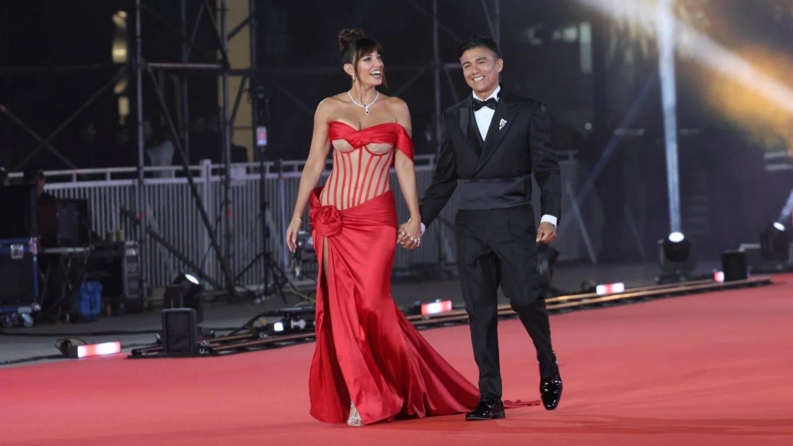 Yamila Reyna con un vestido rojo semitransparente, tomada de la mano de Américo quien usa un traje de dos piezas negro, ambos caminando por una alfombra roja.