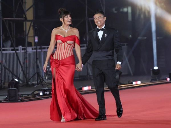 Yamila Reyna con un vestido rojo semitransparente, tomada de la mano de Américo quien usa un traje de dos piezas negro, ambos caminando por una alfombra roja.